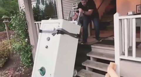a mover guides a tall white gun safe down exterior house steps on a stair-climbing dolly, face visible