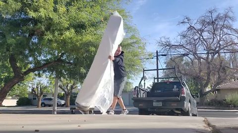 man carrying a plastic-wrapped mattress across a residential street, face visible