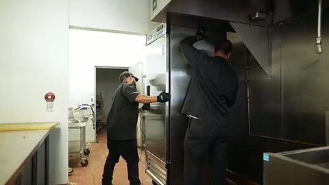two movers rolling a large commercial reach-in refrigerator through a restaurant kitchen, face visible